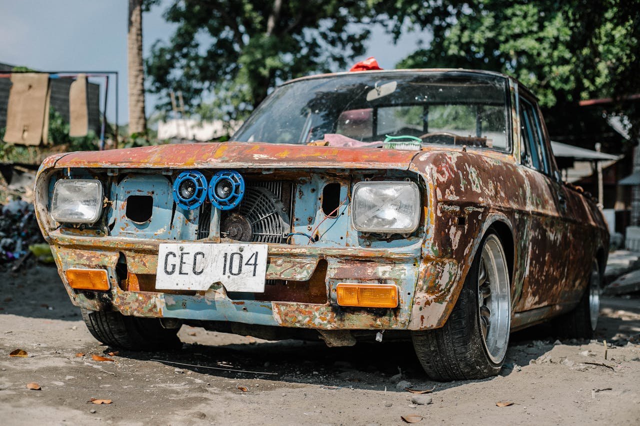 about-us Aged and rusty vintage car parked outdoors on a sunny day, featuring peeling paint and visible wear.